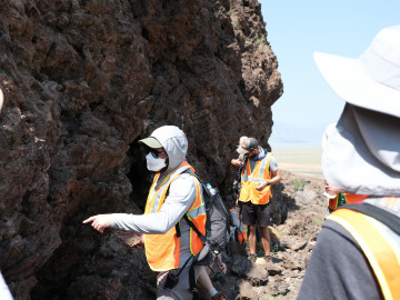 Students in safety vests, hats, and masks look at rock features in a cliff side on a sunny day. Image by Harry Tang