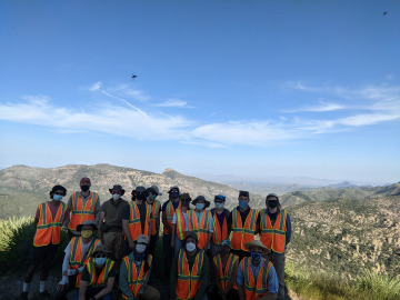 The group of 17 people poses for a picture in their vests in front of a valley with mountains in the back. Some small bugs fly in front of the camera. Image by Robert Melikyan