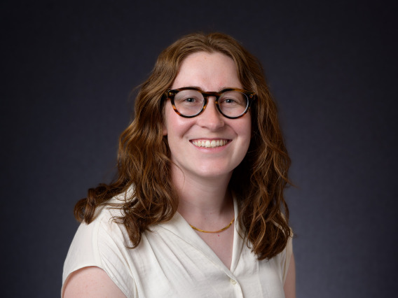 Sophia smiling at camera against dark background. She has shoulder-length wavy brown hair, glasses and freckles.