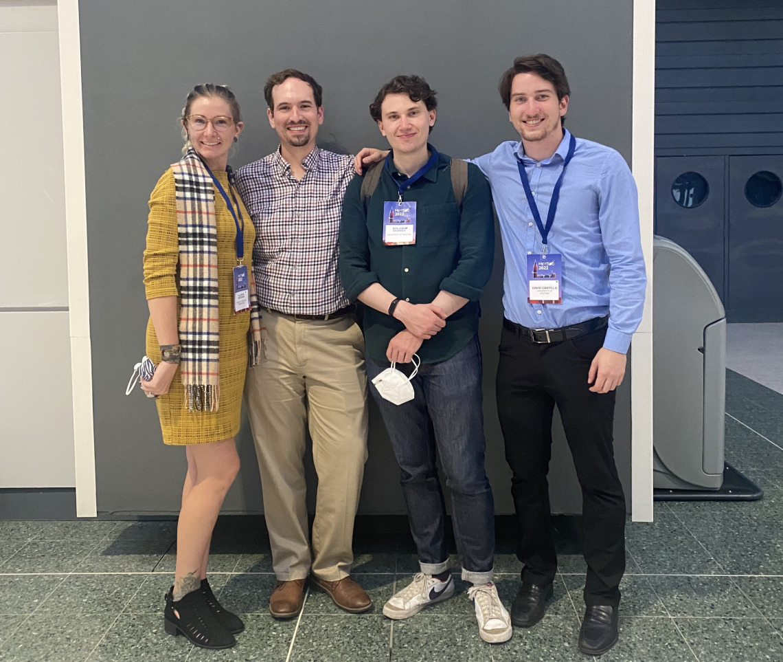 A group of four PhD students posing and smiling for a photo. They are dressed nicely and are standing in front of a plain background.