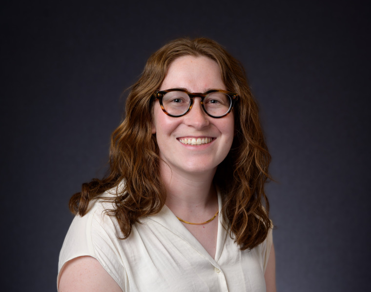 Sophia smiling at camera against dark background. She has shoulder-length wavy brown hair, glasses and freckles.