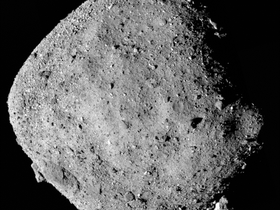 NASA image of a dark, grey asteroid, Bennu. Roughly diamond shaped, it is illuminated from the left with boulders visible on the surface and a large, sloping crater near the dark shadowed edge to the right.